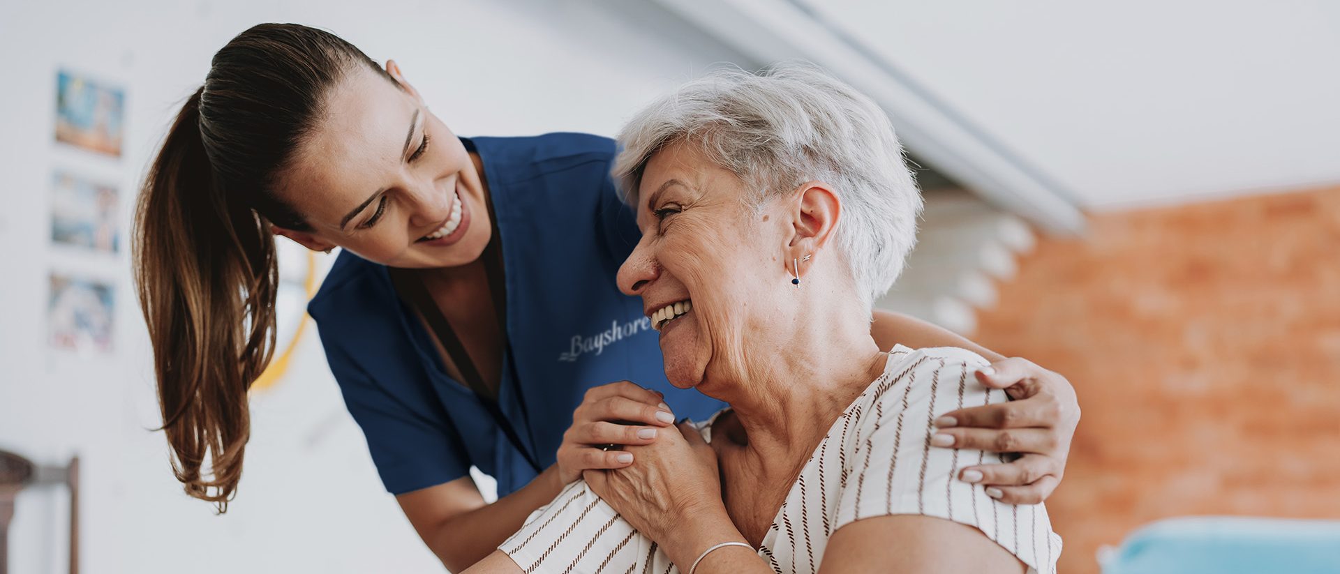 A caregiver and and an elderly women sitting in a chair laugh together