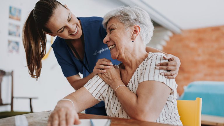 A caregiver and and an elderly women sitting in a chair laugh together